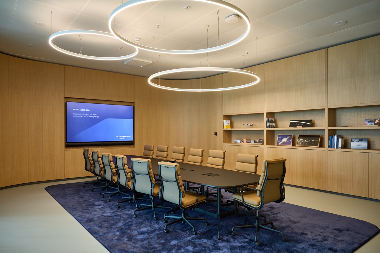 Conference room with a meeting table on carpet, leather chairs, a large screen, a wall unit, and ring-shaped ceiling lights