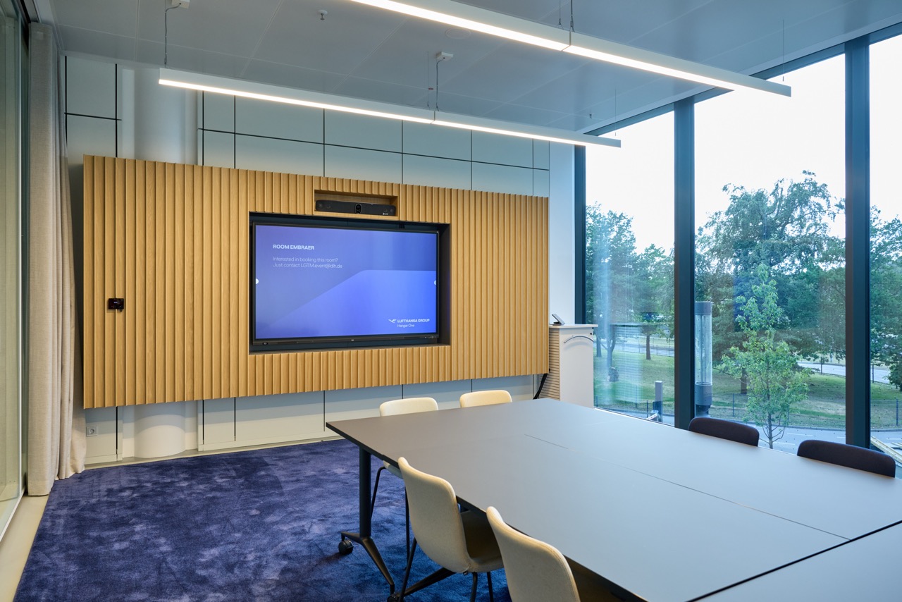 Conference room with a table and chairs on carpet, a large screen, and floor-to-ceiling windows overlooking greenery