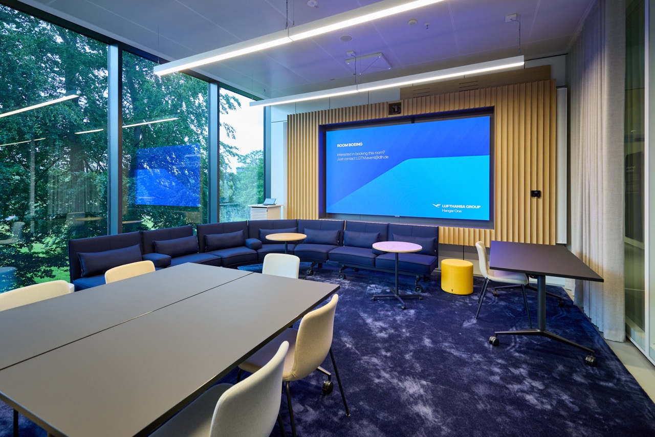 A lounge-style event space with carpet, sofas, tables, a large screen, and floor-to-ceiling windows