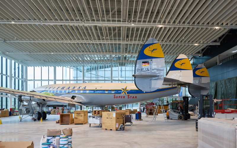 Ein restauriertes historisches Lufthansa-Propellerflugzeug in einer hellen Museumshalle, mit sichtbaren Leitwerken sowie Holzkisten, Leitern und Wartungsausrüstung, die während Aufbau oder Restaurierung darum angeordnet sind.